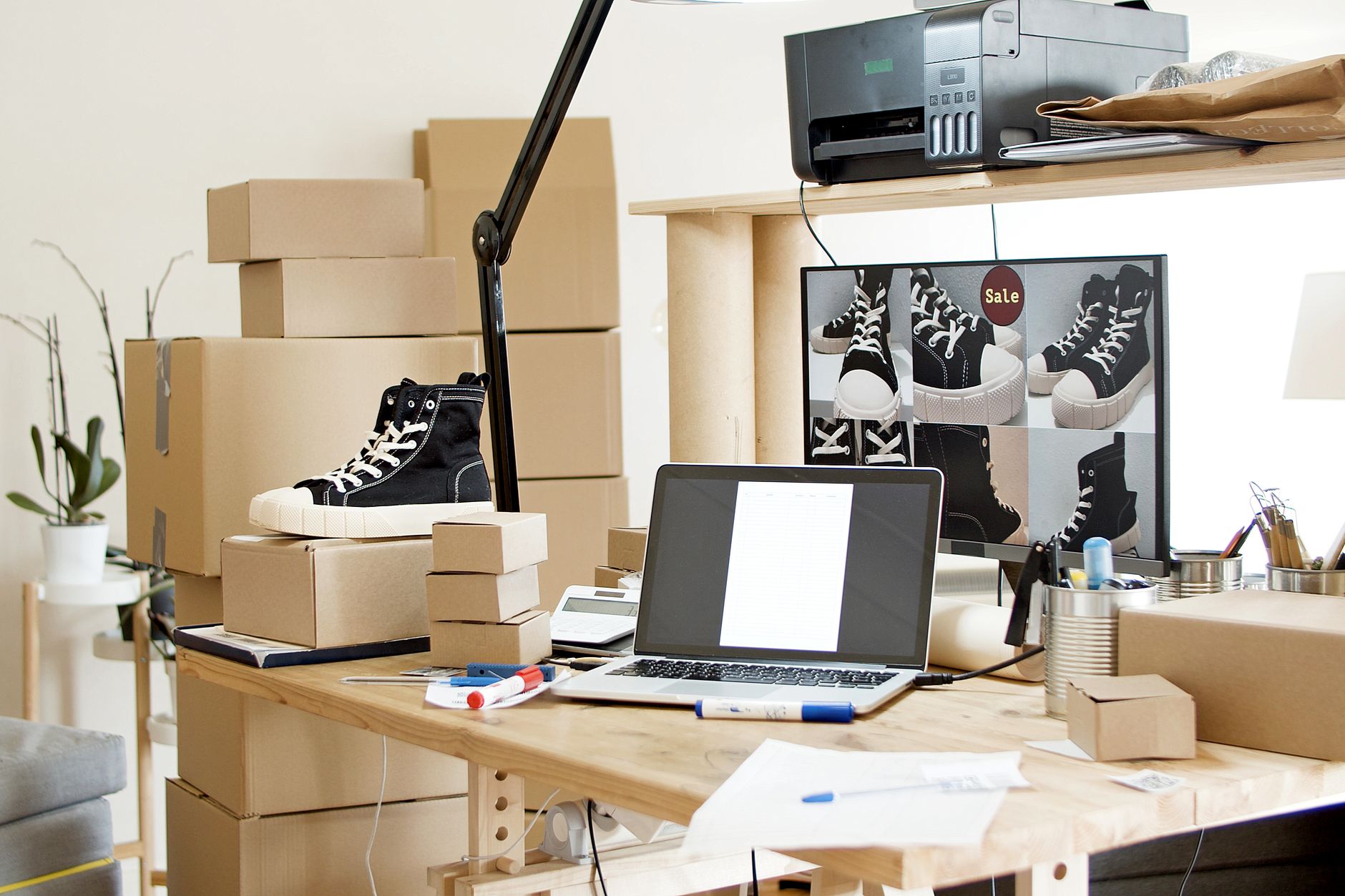 Espace de travail chargé avec chaussures, cartons et ordinateur portable, évoquant une petite activité en ligne gérée seule.