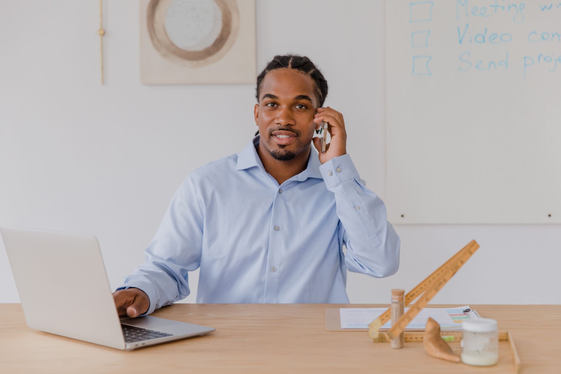 Jeune homme souriant à son bureau, téléphone à la main et ordinateur portable ouvert devant lui.